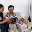 A caregiver talks with a patient while showing her information on a tablet representing the type of thorough communication needed for effective and professional care of home health care patients.